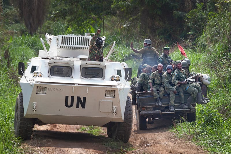 A white MONUSCO armored personnel carrier marked 'UN' is stopped on a dirt road while FARDC soldiers in military uniforms sit on the back of a truck beside it in a green forested area.