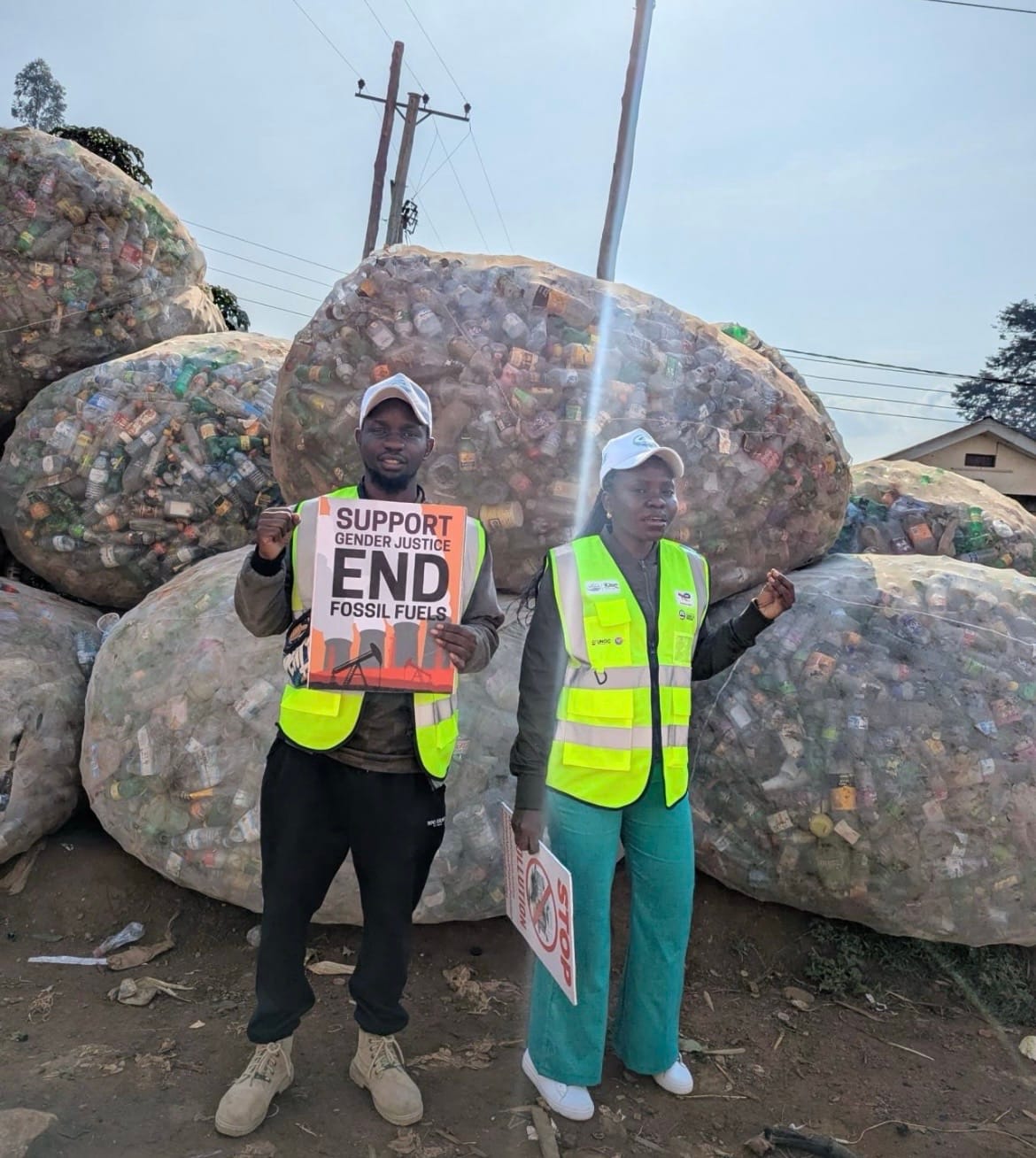 Two people wearing yellow safety vests and caps stand in front of large bags filled with plastic bottles one holds a sign reading 'SUPPORT GENDER JUSTICE END FOSSIL FUELS'.