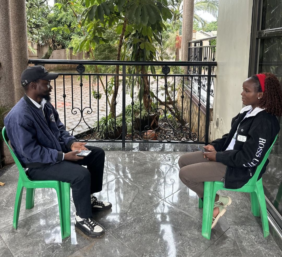 Ali (left) and Clare (right) sitting on green plastic chairs facing each other on a tiled patio with a black metal railing and plants in the background.