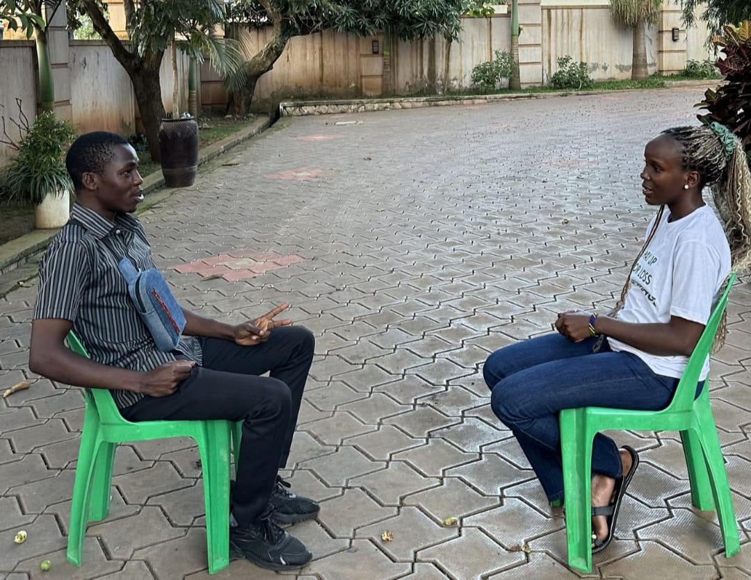 Geofrey (left) and Clare (right) sitting on green plastic chairs facing each other on a paved outdoor area with a wall and plants in the background.
