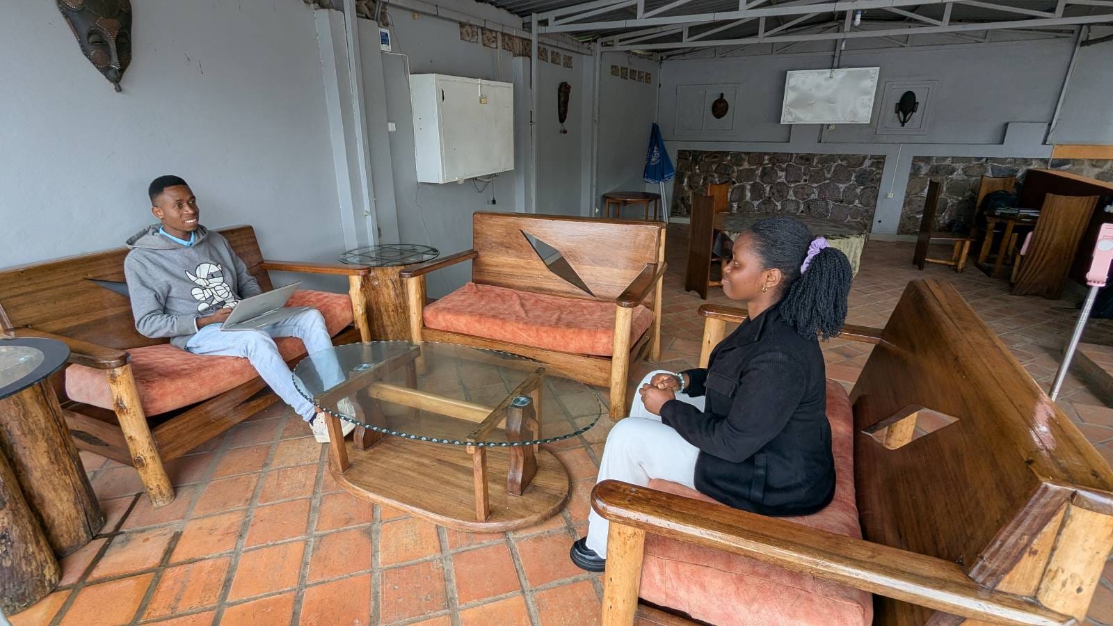 Two people sitting on wooden benches with cushions around a glass coffee table in a room with tiled floor and a stone wall.