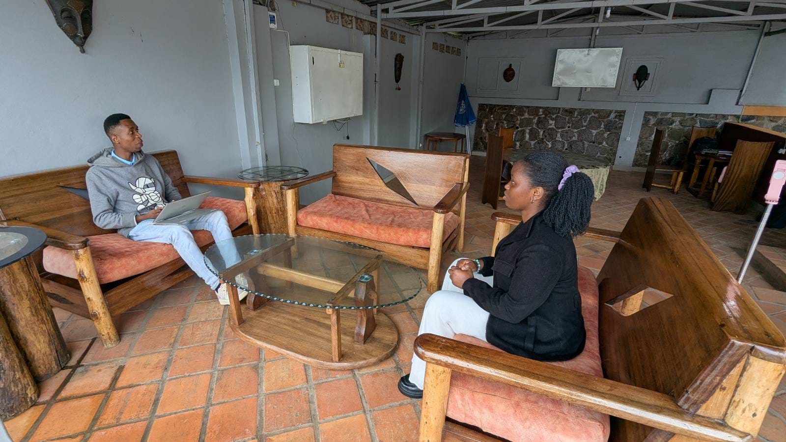 Two people sitting on wooden benches with cushions around a glass coffee table in a room with tiled floor and a stone wall.