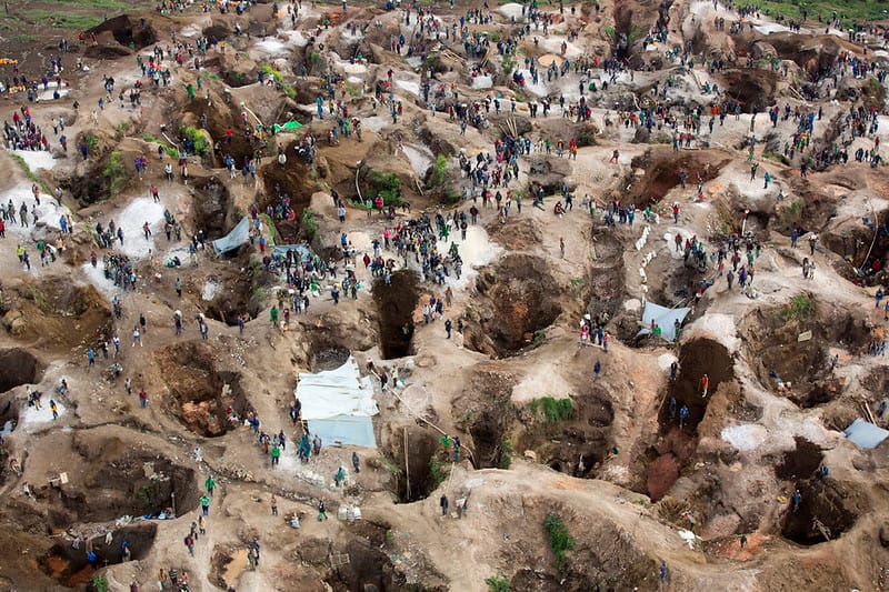 Areal view of a large open-pit mine with numerous irregular pits and many people scattered across the area near makeshift shelters.